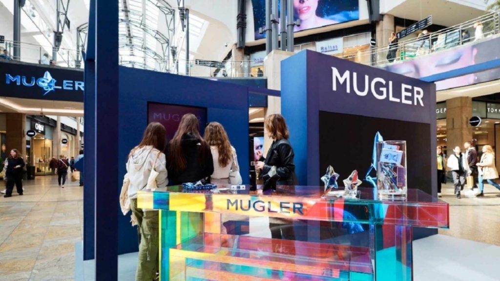 Mugler interactive retail display in a busy shopping mall, showcasing an iridescent counter and perfume samples with engaging digital screens for customer interaction.