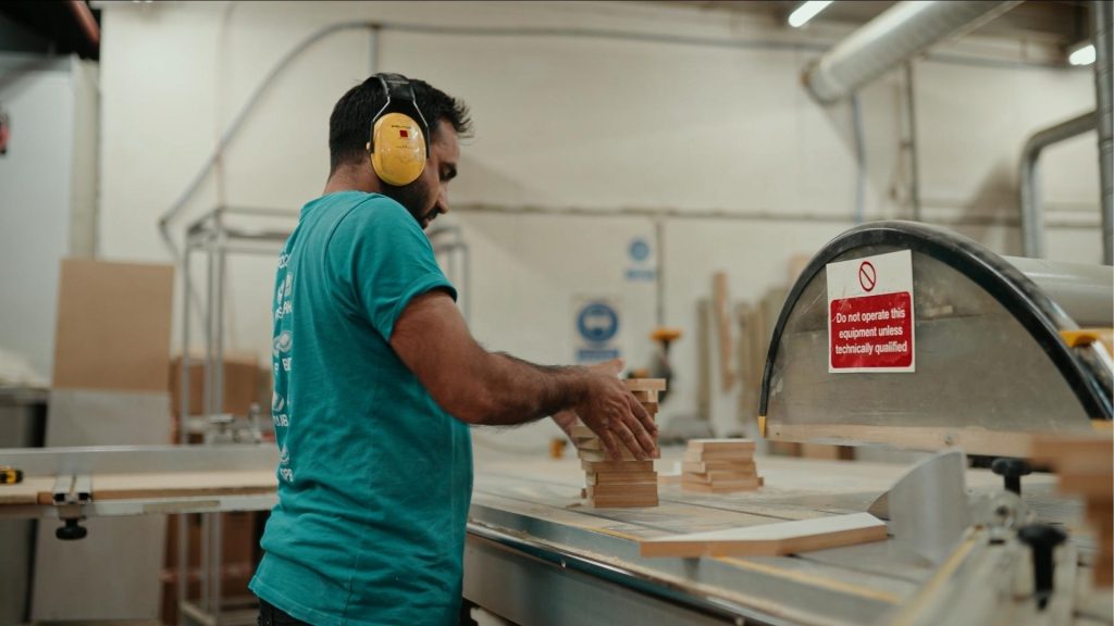 A workshop technician wearing ear protection operates machinery while handling stacked timber components at Ripple’s manufacturing facility.